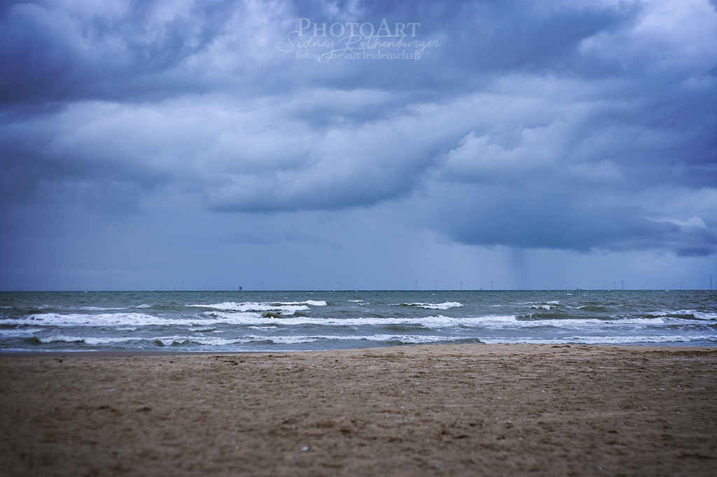 Wildes Meer von Scheveningen. Dank kleiner Lichttemperaturspielerei ist das Bild überwiegend blau. Im Hintergrund sieht man einen Sturm und Regen.