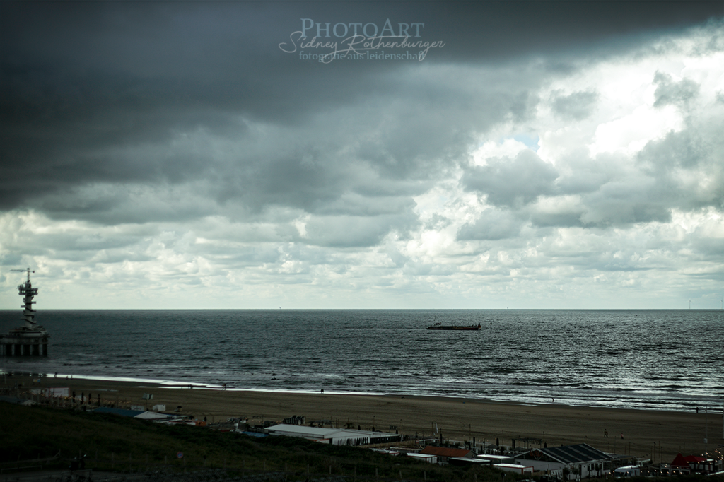 Ein Frachter auf der Nordsee. Links im Bild der Ausläufer von De Pier mit dem Bungeeturm. Die Sonne bricht durch schwere Wolken.
