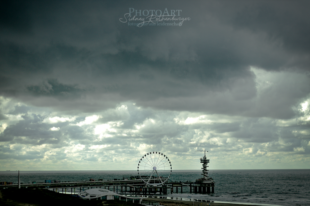 De Pier von Scheveningen/Den Haag. Zu sehen sind das Riesenrad und der Bungeeturm. Im Hintergrund scheint die Sonne durch schwere Wolken.